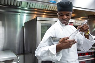 A student in a chef's jacket and black hat whisking holding a silver bowl.